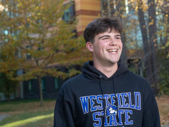 Student wearing black WSU sweatshirt with blue writing and owl logo smiling with fall trees in background.