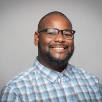 Joseph Seal, Academic Support Coordinator for the Course Achievement, Retention, and Engagement Center. He wears a white and blue-checkered button-down shirt and black glasses. He smiles in front of a plain, gray background.