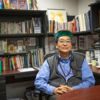 Professor Max Saito of the Communication Department. He sits in his office, which is walled with several shelves of books. His hands are clasped over his torso, and he smiles at the camera in a vest and green hat.
