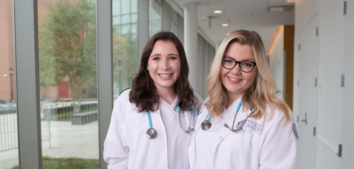 Two nursing students wearing white scrubs and stethoscopes smile at the camera.