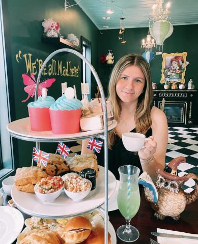 Westfield State student Madeline LeBlanc holds a tea cup and looks out from behind a tray of sandwiches and desserts in a restaurant decorated with an Alice in Wonderland theme.