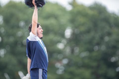 A male cheerleader dressed in navy blue throws one hand into the air while he cheers.