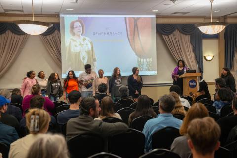 A crowd full of people sit in front of a presentation titled: The Voice of Resilience. One of the presenters stands in front of a podium and addresses the crowd.