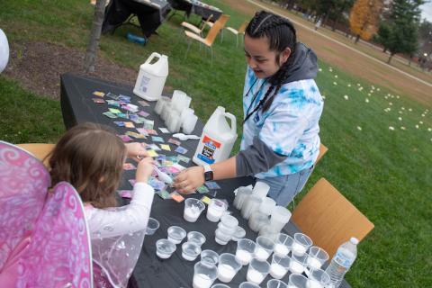 A student in a tie-die sweater makes arts and crafts outside with a young girl in a butterfly costume at a table.