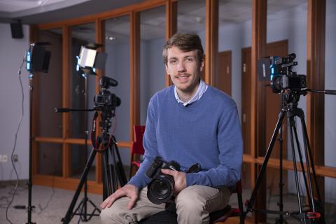 Nathan Dion, class of 2023, sits among a three professional cameras that he uses at John Garvey Inc., as their new Digital Public Relations Analyst. He wears a long-sleeved, blue sweater and tan pants. He holds a black camera in his lap while posing on a stool.