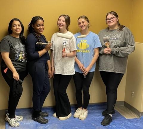 The five students who created a new pediatric waiting room at the MiraVista Behavioral Health Center in Holyoke, MA. Four young women pose in front of a yellow wall, and they're holding paint brushes to show some of the work they've done to create the room.