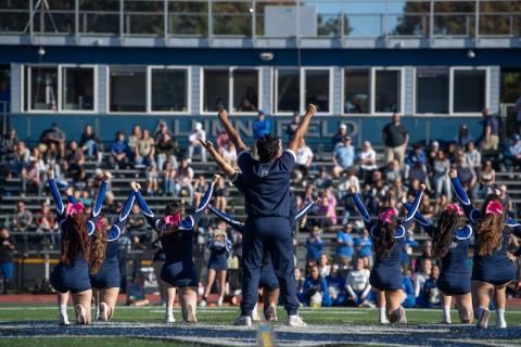 The University's cheerleading team performing in front of a crowd for this year's Homecoming weekend. Owls in blue uniforms face the crowd on bleachers and raise their hands in the air.