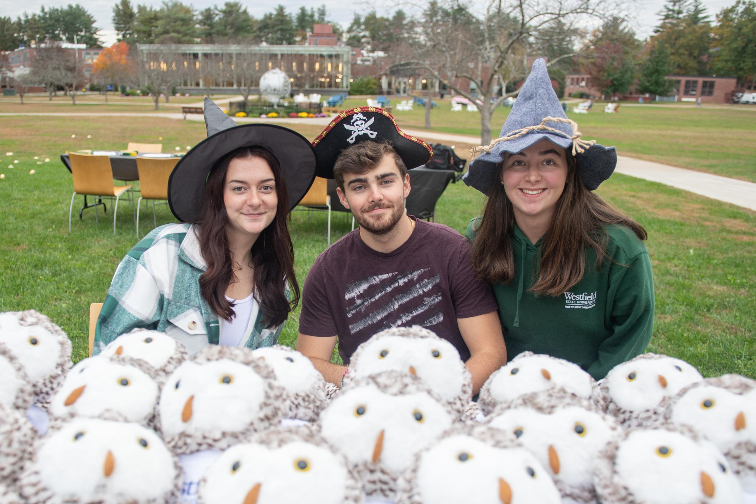 two female and one male Westfield State Students wear silly hats for Halloween as they sit infant of a table of stuffed owls.