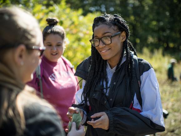 Smiling female student in dark-rimmed glasses talks to her fellow students as they chart the growth of invasive species in a field