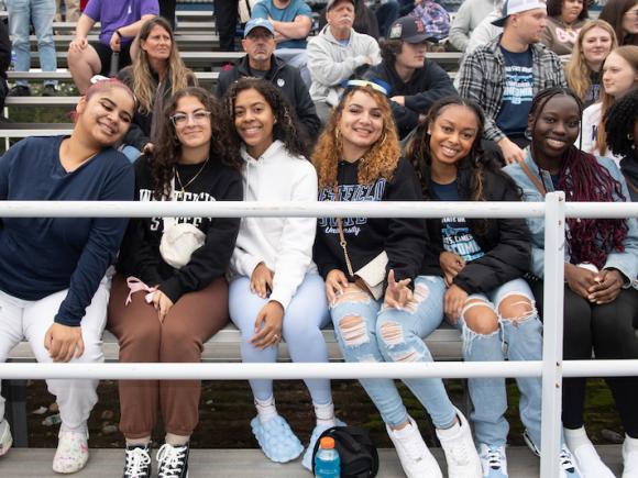 A group of women-students sit on the bleachers and pose next to each other while they cheer on the sports teams.