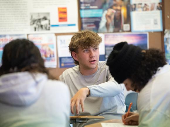 Three Westfield State University students sit in a group of desks while discussing and working on classwork.