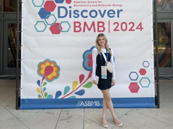 Taylor Camossi at the 2024 BMB Biochemistry Conference in San Antonio. She poses in front of a large white, pink, and blue poster which says, "Discover BMB 2024". She is wearing a white and black dress, white shoes, and a blue lanyard. Graphics of flowers and cells dot the poster behind her.