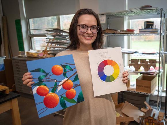 A student holds up a painting of oranges and a color wheel while standing in front of shelves holding many sculptures and other artwork-in-process.
