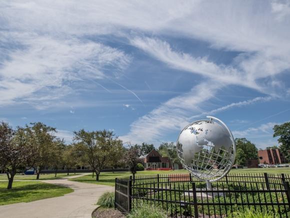The campus globe amidst a sunny, summer day. The sky is blue and cloudy, and trees and grass dot the campus around it.