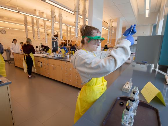 Student in chemistry class wearing a yellow lab apron and eye protection holding a sample.