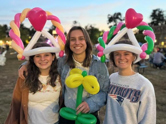 Three women students from the 2024 Family Weekend celebrations. Two are wearing elaborate balloon-art crowns on their heads while another holds a balloon-art green and yellow flower.