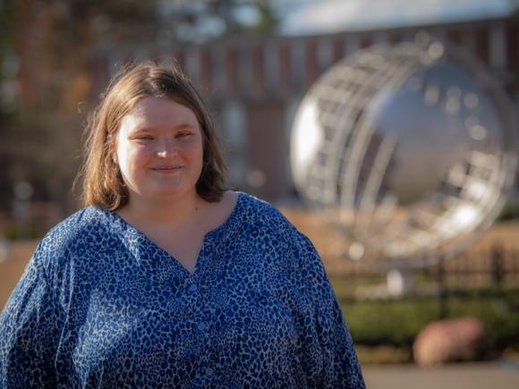 Melissa Costello, Class of 2022, posing in front of the campus globe. She's wearing a blue blouse and has the globe unfocused in the backround.