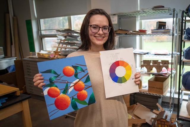A student holds up a painting of oranges and a color wheel while standing in front of shelves holding many sculptures and other artwork-in-process.