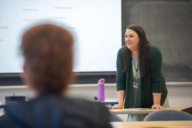 A professor leans on a classroom table and smiles while teaching a history course.