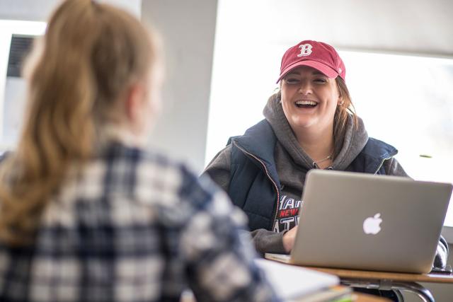 Two Westfield State University students smile and laugh while working on laptops in a classroom.