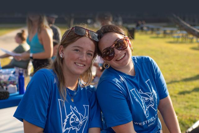 Two female students in blue Westfield State shirts, smile at the camera as they sit on the Campus Green