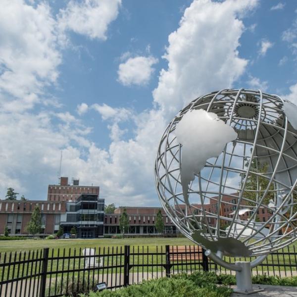 The Westfield State campus globe rests in front of a blue, white-clouded sky. Several brick buildings can be seen in the far background.