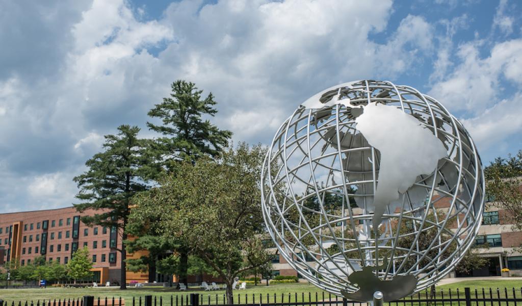 A photo of the campus globe during the summer season. The sky above is blue with clouds, and University Hall can be seen behind the globe itself.