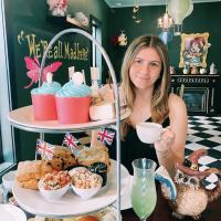 Westfield State student Madeline LeBlanc holds a tea cup and looks out from behind a tray of sandwiches and desserts in a restaurant decorated with an Alice in Wonderland theme.