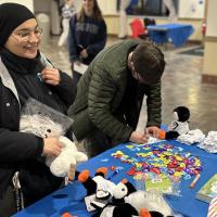 Two students stand in front of a table decorated with black and white penguin plushies, stickers, white socks, and heart-shaped pins. This is for Winter Weekend on campus, where students could make their own winter survival kits for the cold weather.