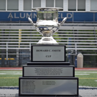 A close-up photo of the Howard C. Smith Cup trophy. It's a large silver chalice with three tiers of printing on its lower levels. It sits on the football field at the University.