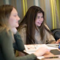 A campus stock photo of two students studying on campus. They have laptops and notebooks on their desk and are laughing at something on the screen.