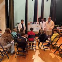 Westfield State Physician Assistant students teach younger students from Westfield Intermediate School. Three PA students in white lab coats present a poster to several younger students who sit in chairs before them.